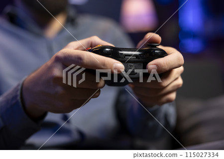Focus shot on controller held by gamer in blurry background playing videogames. Close up shot of gamepad used by man enjoying gaming session in living room while sitting down in front of TV Focus shot on controller held by gamer in blurry background playing videogames. Close up shot of gamepad used by man enjoying gaming session in living room while sitting down in front of TV 115027334