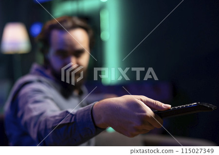 Focus on remote control used by man in blurry background switching channels on television screen. Close up of device used by person to scroll through streaming content, picking movie 115027349