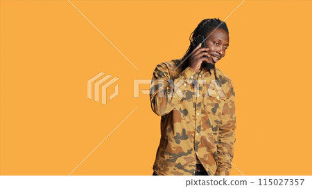 Young adult chatting on phone call in studio, talking to people on remote telephone line over orange background. African american person having fun with telecommunication conversation. Young adult chatting on phone call in studio, talking to people on remote telephone line over orange background. African american person having fun with telecommunication conversation. 115027357