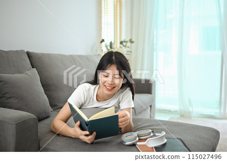 Carefree young asian woman in casual clothes lying on couch and reading book 115027496
