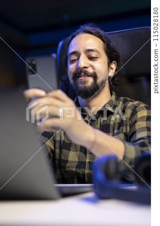 Joyful man holding his smartphone and having an online conversation with friends and family. Thrilled guy using his cell phone to browse the internet and find humorous posts on social media. Joyful man holding his smartphone and having an online conversation with friends and family. Thrilled guy using his cell phone to browse the internet and find humorous posts on social media. 115028180