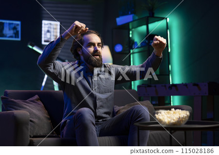 Overjoyed supporter watching sports match on live television broadcast, pleased by team winning trophy. Ecstatic man looking at football competition, raising arms in excitement seeing goal scored 115028186