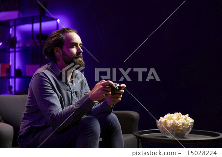 Gamer engaging in online competitive multiplayer tourney using cutting edge gaming console and gamepad while eating popcorn. Man in home theatre playing videogames and consuming snacks 115028224