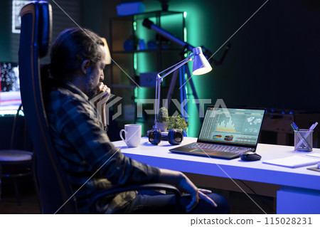 Dedicated man conducting financial research on his laptop, and coming up with ideas. Male individual seated at desk, focused, and productive, uses technology and a notebook for work and study. 115028231