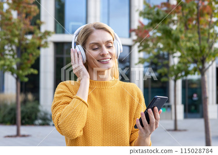 A joyful middle-aged woman in a yellow sweater listens to music on headphones while holding a smartphone, outside by a modern building. A joyful middle-aged woman in a yellow sweater listens to music on headphones while holding a smartphone, outside by a modern building. 115028784