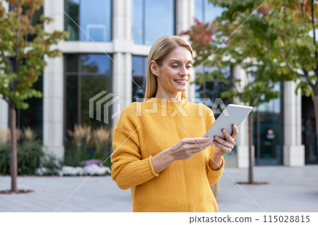 A professional businesswoman in a yellow sweater uses a digital tablet outside a modern office building. She looks content and focused. 115028815