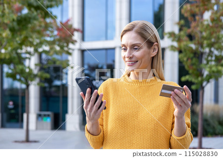 Cheerful blonde woman in yellow sweater using a smartphone and holding a credit card outdoors, possibly shopping or managing finances online. Cheerful blonde woman in yellow sweater using a smartphone and holding a credit card outdoors, possibly shopping or managing finances online. 115028830