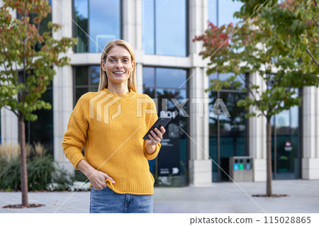 A cheerful woman in a yellow sweater is using her smartphone while standing outdoors near a contemporary office building, enjoying a sunny day. 115028865