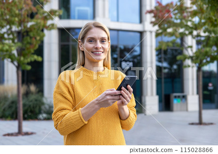 A cheerful woman in a yellow sweater uses her smartphone on a sunny day, with modern buildings forming a soft backdrop. She portrays a relaxed and content demeanor. A cheerful woman in a yellow sweater uses her smartphone on a sunny day, with modern buildings forming a soft backdrop. She portrays a relaxed and content demeanor. 115028866