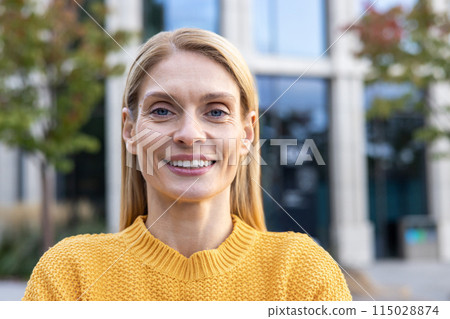 A portrait of a confident middle-aged businesswoman smiling outdoors. She is wearing a yellow sweater, with a modern office building in the background. A portrait of a confident middle-aged businesswoman smiling outdoors. She is wearing a yellow sweater, with a modern office building in the background. 115028874