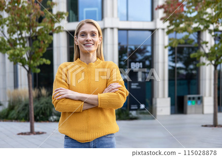 A professional businesswoman stands confidently outdoors, arms crossed, wearing a vibrant yellow sweater. She smiles in a modern urban park setting. 115028878