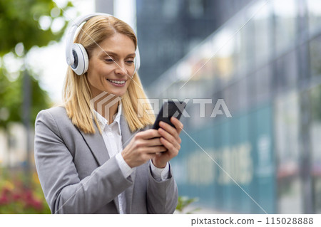 In an urban setting, a modern and professional blonde businesswoman in a grey blazer smiles while using wireless headphones and a smartphone outdoors with city buildings in the background 115028888