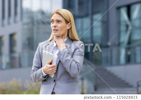 Businesswoman in a gray suit clutching her throat in discomfort while holding a tablet outside an office building. 115028915