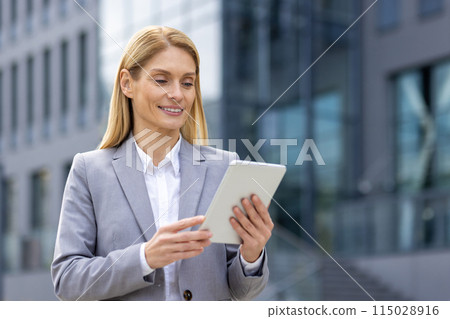 A successful professional businesswoman in a gray suit smiles while using a tablet in a modern urban setting, representing empowerment in the corporate world. She exudes confidence and competence 115028916