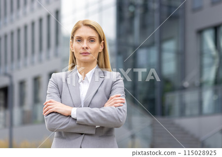 Confident businesswoman in a grey suit standing with her arms crossed outside a modern office building. 115028925
