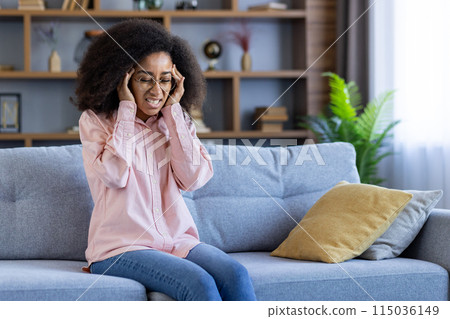 A woman sitting on a couch at home, holding her head in pain and discomfort. She appears distressed while being in a domestic setting. 115036149