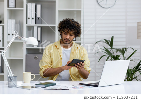 Young professional working at desk in modern office, using a laptop and calculator, surrounded by paperwork. Focused on managing business tasks. 115037211