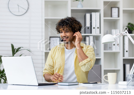 Smiling man wearing a headset while working on a laptop in a modern office setting. Professional workspace with office supplies and plants. 115037213