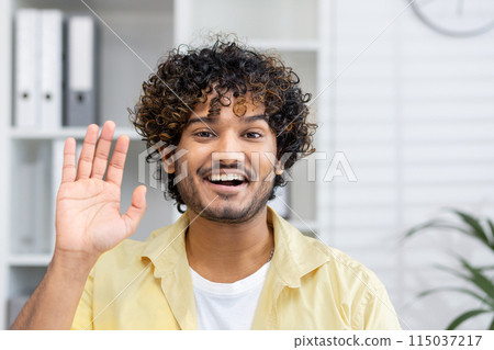 Smiling young man waving during a video call. Friendly and happy interaction in a home office setting. 115037217