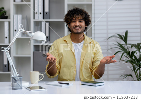 A young man engaging in a video call from his office workspace, smiling and gesturing with hands, portraying a casual and confident communication. 115037218