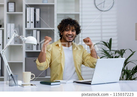A joyful man in a yellow shirt celebrating with raised fists while working on a laptop in his home office. Concept of success, achievement, and positive energy. 115037223
