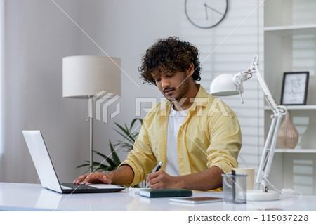 Man in a yellow shirt working remotely from home, using a laptop and taking notes in a notepad at a modern desk. Man in a yellow shirt working remotely from home, using a laptop and taking notes in a notepad at a modern desk. 115037228