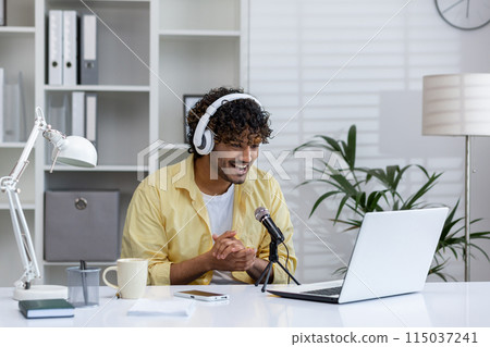 A smiling young man wearing headphones recording a podcast in a modern office setting with a laptop and microphone. 115037241