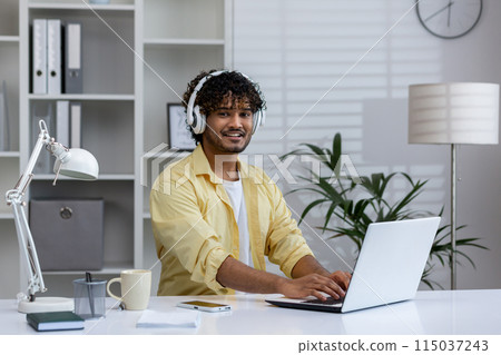 Smiling young man in headphones working on laptop at modern home office. He enjoys a comfortable work environment with stylish decor and natural light. 115037243