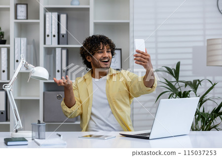 Young man in a modern office taking a selfie with his smartphone, expressing joy and confidence while working at his desk with a laptop. Young man in a modern office taking a selfie with his smartphone, expressing joy and confidence while working at his desk with a laptop. 115037253