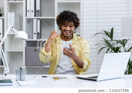 Excited young man celebrating success while looking at smartphone in a modern office setting. Achieving goals and experiencing happiness at work. Excited young man celebrating success while looking at smartphone in a modern office setting. Achieving goals and experiencing happiness at work. 115037266