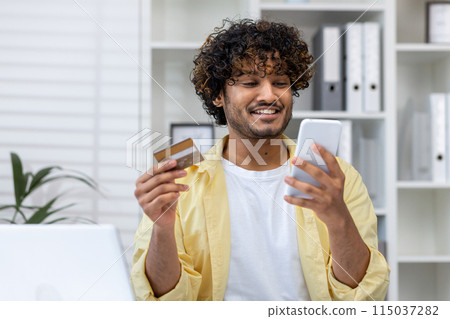 Happy young man using a credit card for online shopping on his smartphone in a modern office setting 115037282