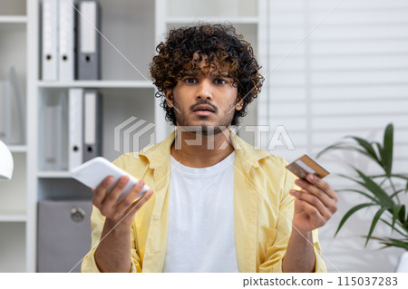 A confused man holding a mobile phone and credit card, looking perplexed while standing in an office environment with shelves and files in the background. A confused man holding a mobile phone and credit card, looking perplexed while standing in an office environment with shelves and files in the background. 115037283