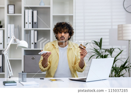 Confused man holding a credit card and smartphone while sitting at an office desk with a laptop, displaying uncertainty. Confused man holding a credit card and smartphone while sitting at an office desk with a laptop, displaying uncertainty. 115037285