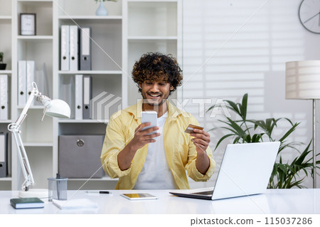 Happy young man using smartphone and credit card for online shopping, sitting in modern home office with laptop and plants. Happy young man using smartphone and credit card for online shopping, sitting in modern home office with laptop and plants. 115037286