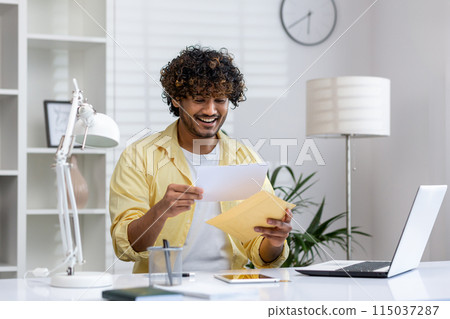 Smiling man opening a letter while sitting at a desk in a modern office. Demonstrates positive emotions, paperwork, and a professional work environment. Smiling man opening a letter while sitting at a desk in a modern office. Demonstrates positive emotions, paperwork, and a professional work environment. 115037287