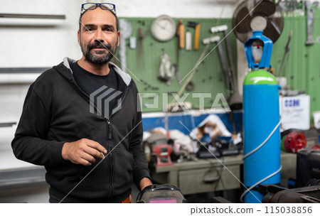 Portrait of a blacksmith during a break in a welding session. He takes the opportunity to smoke a cigarette, the welder's helmet can be seen in the bottom right. Blurred background with workshop. 115038856