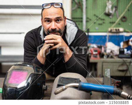 Nice portrait of a Caucasian man looking into camera. On his bench are his welder's work tools Nice portrait of a Caucasian man looking into camera. On his bench are his welder's work tools 115038885