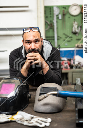 Nice portrait of a Caucasian man looking into camera. On his bench are his welder's work tools Nice portrait of a Caucasian man looking into camera. On his bench are his welder's work tools 115038886