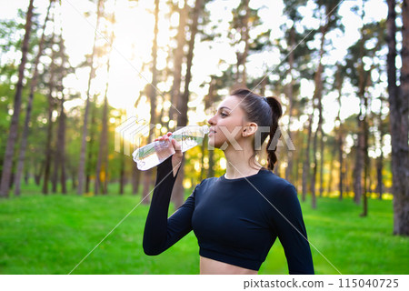 A woman in a sporty black top holds up a water bottle for a drink of water after a sport in the park. A woman in a sporty black top holds up a water bottle for a drink of water after a sport in the park. 115040725