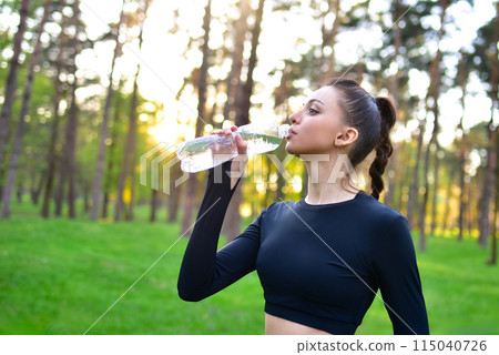 A woman in a sporty black top holds up a water bottle for a drink of water after a sport in the park. 115040726