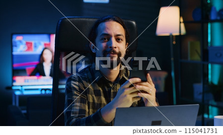 Man relaxing in apartment sitting at office desk, texting friends on cellphone, enjoying leisure time. Smiling remote worker taking break to talk with mates on internet using phone, camera A Man relaxing in apartment sitting at office desk, texting friends on cellphone, enjoying leisure time. Smiling remote worker taking break to talk with mates on internet using phone, camera A 115041731