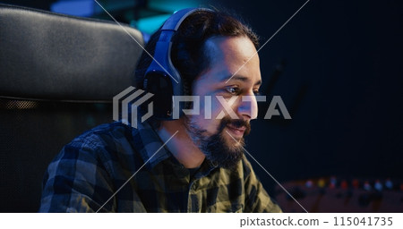 Happy man drinking coffee and writing emails at home office desk while listening music. Smiling remote worker doing b2b networking over internet, wearing headphones, enjoying hot beverage, camera B 115041735