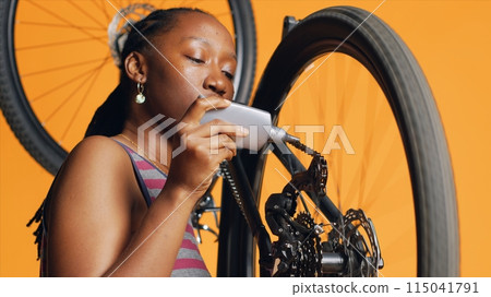 BIPOC mechanic using specialized glue to fix damaged bicycle chain, orange studio background. Expert applying adhesive on bike parts during checkup process, close up shot 115041791