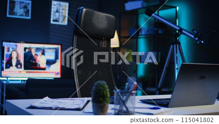 Apartment illuminated by RGB lights with office desk in front of smart TV displaying news program broadcasting. Laptop, mini house plants and headphones on table in empty room, camera B close up shot 115041802