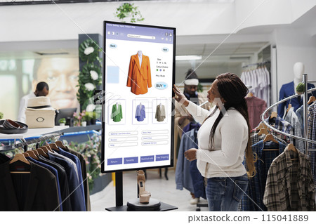 African american client selecting items to buy from self service checkout kiosk, reviewing modern clothes and sizes on interactive monitor. Young woman looking at products on board. 115041889