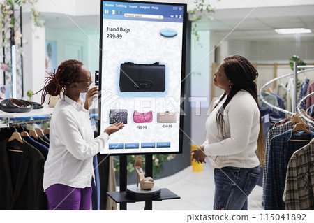 Store employee advising customer with kiosk service, assistant helping client in fashion boutique. Young buyer searching for clothing items on touch screen display board, retail shop. 115041892