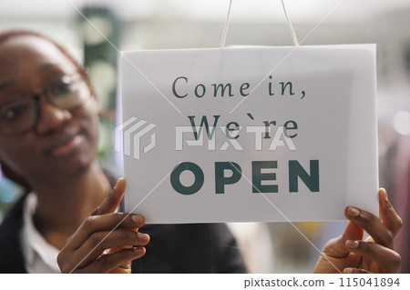 Fashion clothes shop manager holding opening signboard with come in message. Apparel store young african american woman assistant hanging sign informing clients about workday beginning 115041894