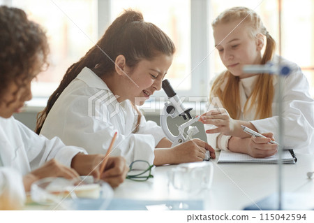 Selective focus shot of Middle Eastern girl wearing lab coat sitting at table in classroom looking through microscope during Chemistry lesson Selective focus shot of Middle Eastern girl wearing lab coat sitting at table in classroom looking through microscope during Chemistry lesson 115042594