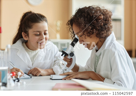 African American boy wearing lab coat sitting in classroom looking through microscope while Middle Eastern girl making notes in notebook African American boy wearing lab coat sitting in classroom looking through microscope while Middle Eastern girl making notes in notebook 115042597