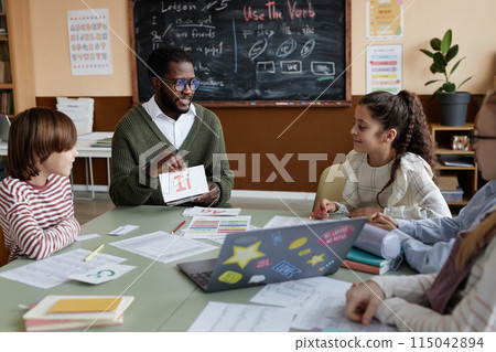 Young African American teacher of English sitting at table in classroom showing alphabet cards to kids during lesson 115042894